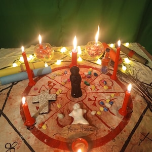 May include: A circular cloth featuring red geometric symbols and a central black figurine, surrounded by six red candles, a white clay figure, a stone cross, small colorful stones, a small glass bottle, and a leaf with the text Your Name written on it.