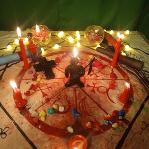 May include: A circular cloth featuring a red occult sigil, centered with a black horned figurine holding a scythe. The arrangement includes a black cross, small colorful stones, two glass bowls, rolled scrolls, and five lit red taper candles.