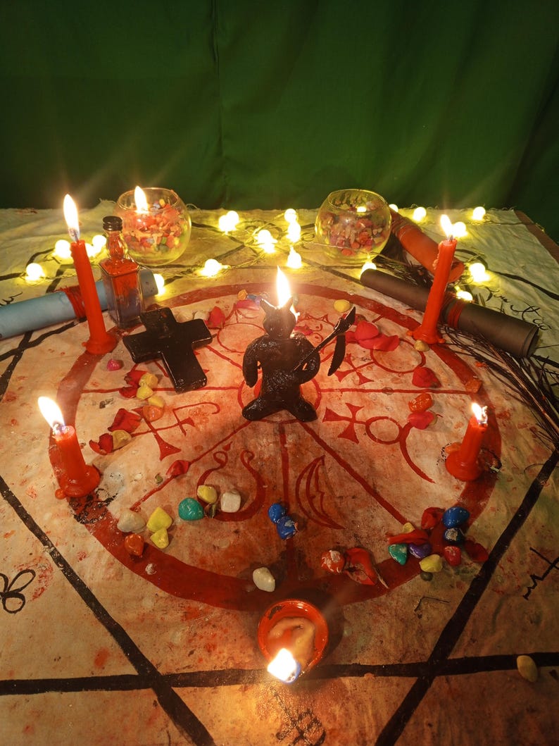 May include: A circular cloth featuring a red occult sigil, centered with a black horned figurine holding a scythe. The arrangement includes a black cross, small colorful stones, two glass bowls, rolled scrolls, and five lit red taper candles.