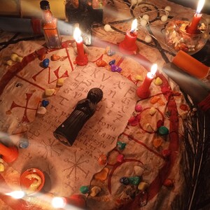 May include: A circular beige cloth featuring red hand-drawn symbols and text, arranged with a black figurine, multiple red candles, small glass bowls containing colorful stones, a black cross, and a small glass bottle with orange liquid.