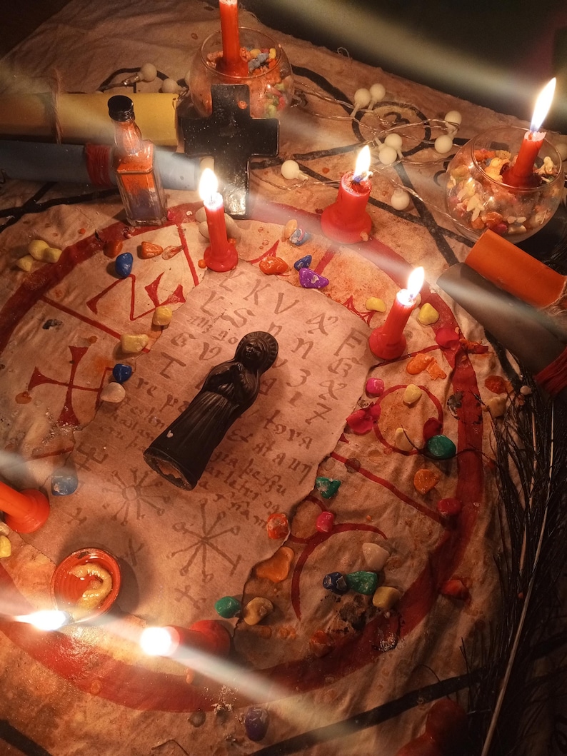 May include: A circular beige cloth featuring red hand-drawn symbols and text, arranged with a black figurine, multiple red candles, small glass bowls containing colorful stones, a black cross, and a small glass bottle with orange liquid.