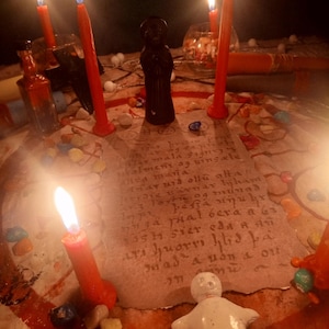 May include: A circular parchment with handwritten text sits at the center of a ritual arrangement featuring five lit red candles, a small black figurine, a white doll, and scattered colorful stones. The scene is illuminated by the warm glow of flames.