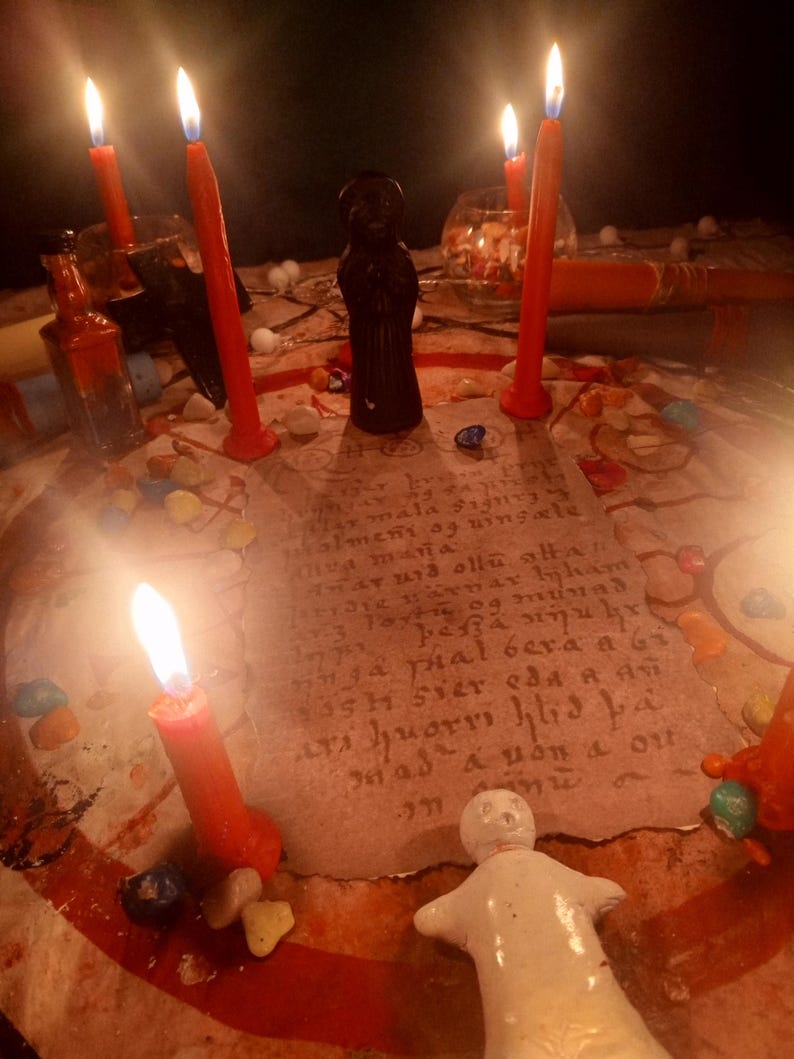 May include: A circular parchment with handwritten text sits at the center of a ritual arrangement featuring five lit red candles, a small black figurine, a white doll, and scattered colorful stones. The scene is illuminated by the warm glow of flames.