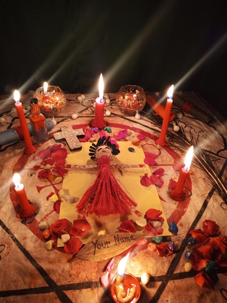 May include: A handcrafted doll with dark hair and a red dress placed on a yellow paper with the text Your Name. The doll is surrounded by six red candles, scattered red rose petals, colorful small stones, and a silver cross on a patterned cloth.