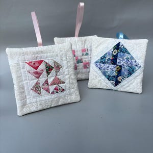 May include: Three square quilted fabric ornaments with white floral patterned backgrounds. Each features a central patchwork design in pink, blue, or multi-coloured floral prints. Each ornament includes a thin satin ribbon loop for hanging.