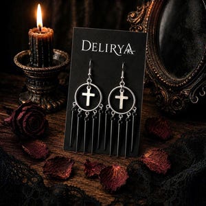 Può includere: Orecchini color argento con un design a croce, appesi a una carta nera con il marchio "DELIRYA". Gli orecchini hanno un design circolare con una croce e barre verticali pendenti. Una candela accesa e petali di rosa sullo sfondo.