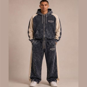 Puede incluir: Un chándal de dos piezas a juego en un lavado azul oscuro con paneles laterales color crema. La chaqueta con capucha y cremallera y los pantalones de pernera ancha llevan el texto "NORIX STUDIO" en blanco. El modelo lleva zapatillas blancas.