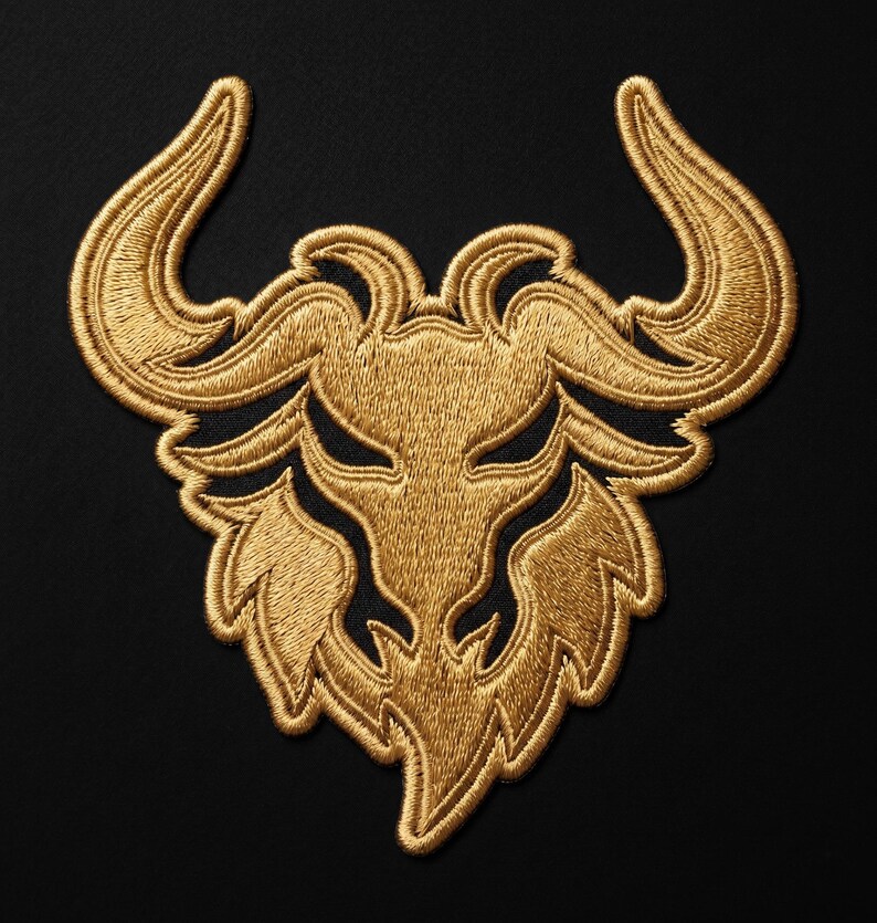 May include: Gold embroidered patch featuring a stylized bull's head with curved horns and a detailed face. The patch is set against a black background, showcasing the intricate threadwork and design.