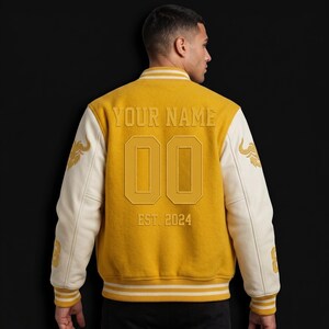 May include: Mustard yellow varsity jacket with cream-colored sleeves. The back features the text "YOUR NAME", the number "00", and "EST. 2024". Gold embroidered details are on the sleeves. The jacket has a ribbed collar and cuffs.