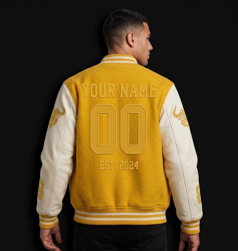 May include: Mustard yellow varsity jacket with cream-colored sleeves. The back features the text "YOUR NAME", the number "00", and "EST. 2024". Gold embroidered details are on the sleeves. The jacket has a ribbed collar and cuffs.
