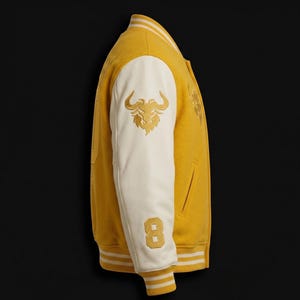 May include: A yellow varsity jacket with cream-colored sleeves. The jacket features a gold embroidered bull logo on the sleeve and the number 8. White and gold striped trim accents the collar, cuffs, and hem. The jacket is displayed against a black background.