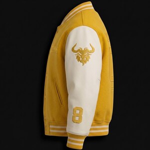 May include: Mustard yellow varsity jacket with white sleeves. Features a gold embroidered bull logo on the sleeve and the number 8. The jacket has a ribbed collar, cuffs, and hem with white and yellow stripes. A classic, sporty design.
