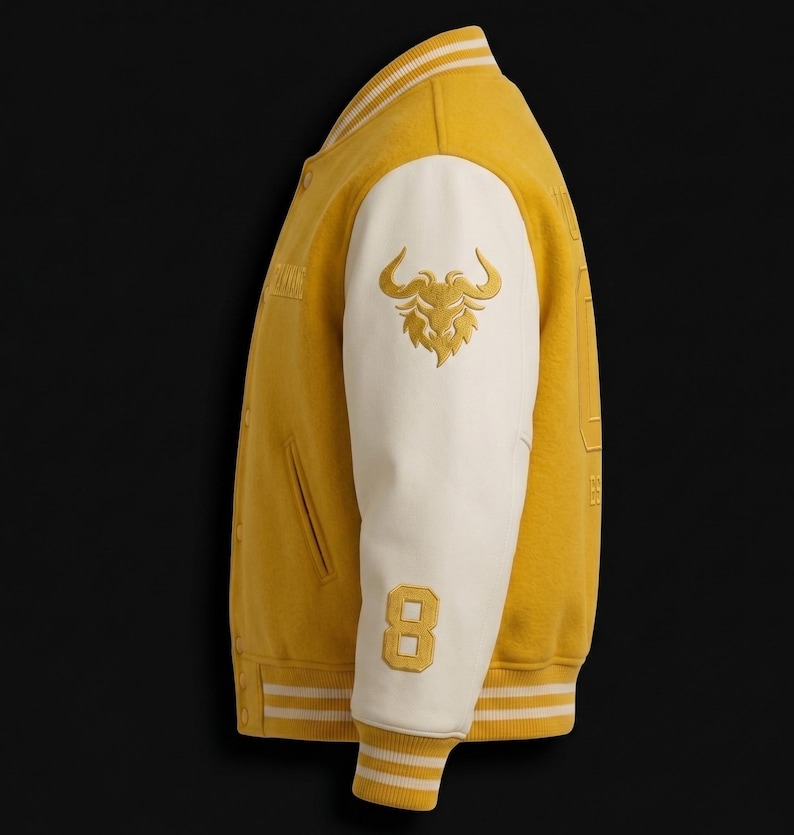 May include: Mustard yellow varsity jacket with white sleeves. Features a gold embroidered bull logo on the sleeve and the number 8. The jacket has a ribbed collar, cuffs, and hem with white and yellow stripes. A classic, sporty design.