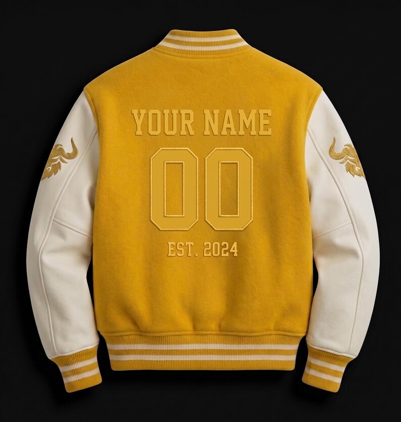 May include: Mustard yellow varsity jacket with cream-colored sleeves. The back features the text "YOUR NAME", the number "00", and "EST. 2024". Gold embroidered details are on the sleeves. The jacket has a ribbed collar, cuffs, and hem with white stripes.
