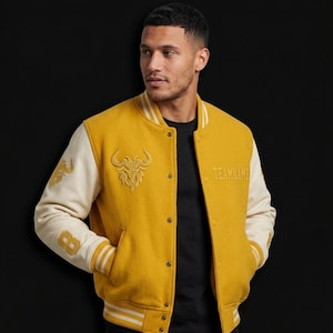 May include: Mustard yellow varsity jacket with cream-colored sleeves. Features a bull graphic and the word "TEAMNAME" embroidered on the front. The left sleeve has the number "8" and a design. The jacket has a button-up closure and striped trim.