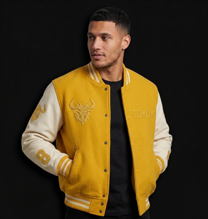 May include: Mustard yellow varsity jacket with cream-colored sleeves. Features a bull graphic and the word "TEAMNAME" embroidered on the front. The left sleeve has the number "8" and a design. The jacket has a button-up closure and striped trim.