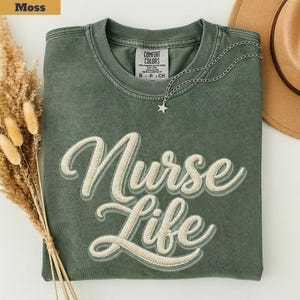 May include: A moss green crewneck t-shirt featuring the embroidered text Nurse Life in cream-colored script. The shirt has a Comfort Colors label at the inner neckline and is paired with a silver-toned star pendant necklace on a delicate chain.