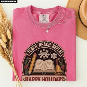 May include: A pink short-sleeved t-shirt featuring a graphic of an open book, a pencil, and a bell surrounded by snowflakes and greenery. The text reads "TEACH. REACH. REPEAT" and "HAPPY HOLIDAYS" in a dark brown arched frame on the front.