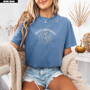 May include: A blue short-sleeved t-shirt featuring a graphic print of a Bigfoot silhouette surrounded by pine trees. The text reads PACIFIC NORTHWEST and CRYPTID SOCIETY. The shirt has a crew neckline and is worn with light blue denim shorts.