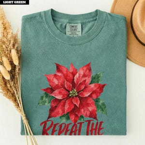 May include: A light green crew neck t-shirt featuring a large red poinsettia flower graphic in the center. Below the flower, the text reads REPEAT THE. The shirt has a Comfort Colors label at the inner neckline and a short-sleeved design.