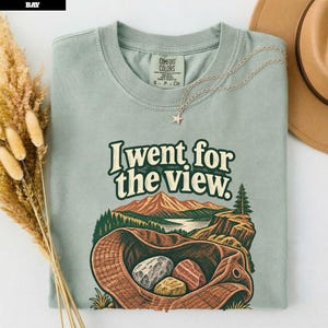 Könnte beinhalten: Ein salbeigrünes T-Shirt mit Rundhalsausschnitt und einem Grafikdruck eines Fischerhuts mit drei Steinen vor einer Landschaft aus Bergen, Bäumen und einem See. Der Text "I went for the view." ist in dunkelgrüner Schrift über dem Motiv gedruckt.