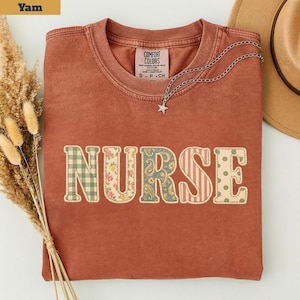 Puede incluir: Una camiseta de manga corta color terracota con la palabra NURSE impresa en letras grandes en el pecho. Cada letra presenta un patrón diferente, incluyendo cuadros vichy verdes, flores, cachemir, rayas rojas y lunares verdes.
