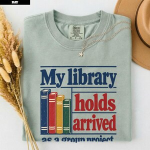 May include: A sage green short-sleeve t-shirt featuring a graphic of four vertical books in blue, red, yellow, and green. The text reads My library holds arrived. The shirt has a Comfort Colors label at the inner neckline and a star-shaped pendant.