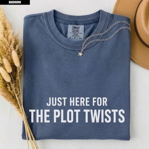 May include: A denim blue short-sleeved t-shirt with a crew neckline and the white text "JUST HERE FOR THE PLOT TWISTS" printed on the front. The shirt features a Comfort Colors brand label at the inner neck and a silver star pendant necklace.