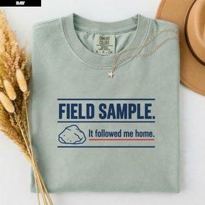 Pode incluir: Uma t-shirt de gola redonda em verde sálvia com o texto azul escuro "FIELD SAMPLE." acima de um gráfico de uma rocha e o texto "It followed me home.". A peça tem uma etiqueta Comfort Colors no decote interno e um colar de estrela prateado.