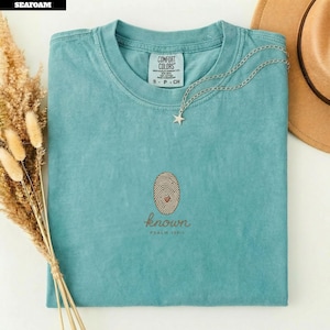 Op de afbeelding: Een zeegroen t-shirt met korte mouwen en een Comfort Colors-label. Op de borst staat een geborduurd ovaal vingerafdrukontwerp met een klein hartje en de tekst known PSALM 139:1. Een zilveren ster-halsketting rust op de halslijn.