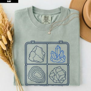 以下が含まれることがあります： セージグリーンの半袖Tシャツ。4種類の鉱物と結晶の標本がグリッド状に配置されたダークブルーのグラフィックプリントが施されています。クルーネックで、首の内側にComfort Colorsのブランドラベルがプリントされています。