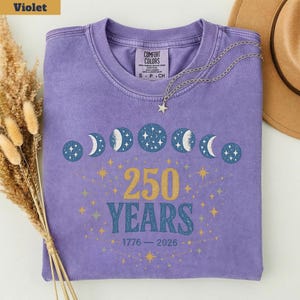 Op de afbeelding: Een violet Comfort Colors T-shirt met een hemels ontwerp van maanfasen en sterren. Het shirt heeft de tekst "250 YEARS" in goud, met de jaren 1776-2026 eronder. Een zilveren ketting met een sterhanger ligt op het shirt.