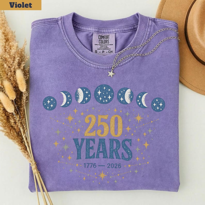 Op de afbeelding: Een violet Comfort Colors T-shirt met een hemels ontwerp van maanfasen en sterren. Het shirt heeft de tekst "250 YEARS" in goud, met de jaren 1776-2026 eronder. Een zilveren ketting met een sterhanger ligt op het shirt.