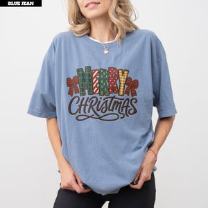 Puede incluir: Una camiseta de manga corta color azul vaquero con el texto MERRY CHRISTMAS en una fuente con varios patrones y lazos rojos a los lados. La palabra MERRY se muestra con diversos motivos festivos, mientras que CHRISTMAS está en letra marrón.