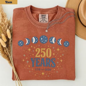 Op de afbeelding: Een roestkleurig T-shirt met een grafisch ontwerp met maanfasen, sterren en de tekst "250 YEARS" met de jaren 1776-2026. Het shirt heeft een Comfort Colors-label en een zilveren ketting met een sterhanger.