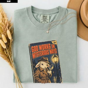 May include: A sage green crew neck t-shirt featuring a graphic print of a lamb dressed as a detective in a trench coat and fedora, holding a magnifying glass under a streetlamp. The text reads God Works In Mysterious Ways with a 25c price tag.