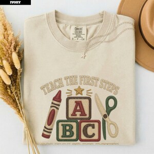 May include: An ivory crewneck t-shirt featuring an embroidered design with the text TEACH THE FIRST STEPS. The graphic includes a red crayon, a gold star, three alphabet blocks labelled A, B, and C, and green scissors. The shirt has a Comfort Colors label.
