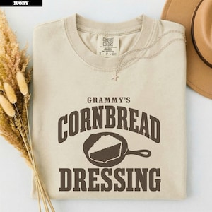 以下が含まれることがあります： アイボリー色の半袖Tシャツ。ダークブラウンの文字で「GRAMMY'S CORNBREAD DRESSING」とプリントされ、その下に鋳鉄製のスキレットに入ったコーンブレッドのイラストが描かれています。クルーネックで、首元にComfort Colorsのタグが付いています。