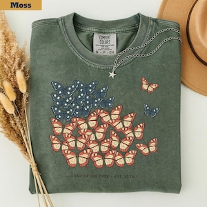 以下が含まれることがあります： モスグリーンのTシャツ。赤、白、青の蝶のデザインで、アメリカの国旗を連想させる。Tシャツには「LAND OF THE FREE - EST. 1976」の文字。星のペンダントが付いたシルバーチェーンが飾られている。