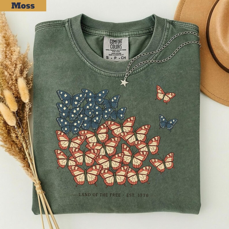以下が含まれることがあります： モスグリーンのTシャツ。赤、白、青の蝶のデザインで、アメリカの国旗を連想させる。Tシャツには「LAND OF THE FREE - EST. 1976」の文字。星のペンダントが付いたシルバーチェーンが飾られている。