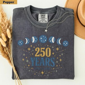 Op de afbeelding: Donkergrijze crewneck sweatshirt met een hemels ontwerp. De afbeelding toont maanfasen, sterren en de tekst "250 YEARS 1776-2026" in goud en blauw. Een zilveren ketting met een sterhanger ligt op de trui.
