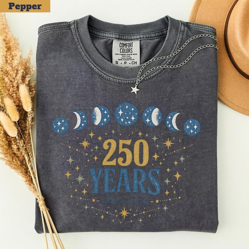 Op de afbeelding: Donkergrijze crewneck sweatshirt met een hemels ontwerp. De afbeelding toont maanfasen, sterren en de tekst "250 YEARS 1776-2026" in goud en blauw. Een zilveren ketting met een sterhanger ligt op de trui.