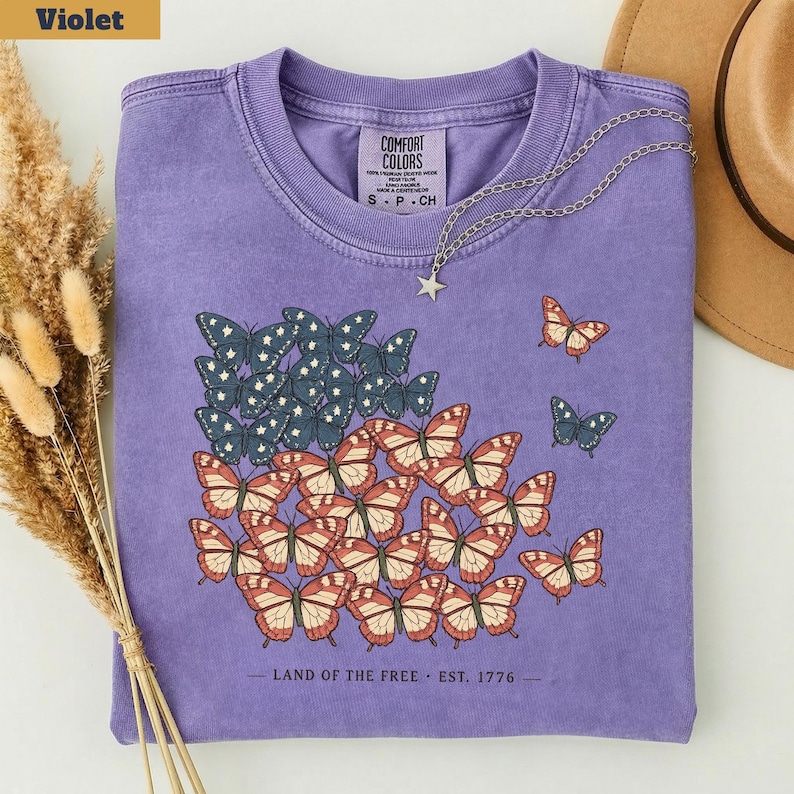 以下が含まれることがあります： アメリカ国旗の形をした蝶のデザインが特徴の、紫色のComfort Colors Tシャツ。赤、白、青の蝶のデザインが含まれています。「LAND OF THE FREE. EST. 1776」の文字が下部にプリントされています。星のペンダントが付いたシルバーのネックレスも確認できます。