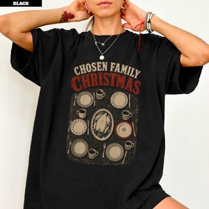 May include: A black t-shirt featuring a graphic print of a holiday dinner table setting. The text reads CHOSEN FAMILY CHRISTMAS in beige and red. The illustration includes plates, mugs, silverware, and a central roasted turkey on a platter.