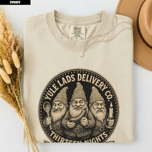 Puede incluir: Una camiseta de color marfil con un gráfico circular negro de tres gnomos barbudos con sombreros puntiagudos. El texto dice YULE LADS DELIVERY CO. y THIRTEEN NIGHTS. Un gnomo sostiene una cuchara de madera, otro una salchicha y otro una vela.