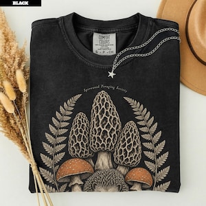 Könnte beinhalten: Ein schwarzes T-Shirt mit Rundhalsausschnitt und einem Grafikdruck von drei Morcheln, zwei Pilzen mit orangefarbenen Kappen und einem Igel, umgeben von Farnwedeln. Der Text lautet Sporewood Foraging Society. Eine silberne Stern-Halskette liegt auf dem Kragen.