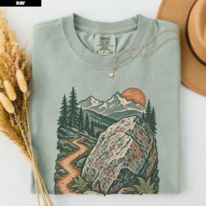 以下が含まれることがあります： セージグリーンの半袖Tシャツ。山岳風景、曲がりくねった小道、大きな岩、松の木、太陽のグラフィックプリントが施されています。クルーネックで、襟の内側にComfort Colorsのブランドラベルがプリントされています。