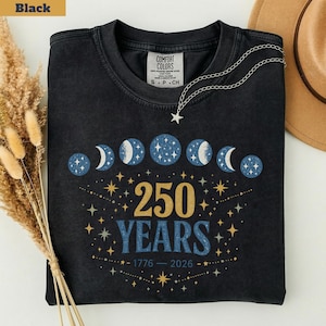 Op de afbeelding: Zwart T-shirt met een hemels ontwerp met maanfasen en sterren. Het shirt toont de tekst "250 YEARS" met de jaren 1776-2026. Een zilveren ketting met een sterhanger ligt op het shirt.