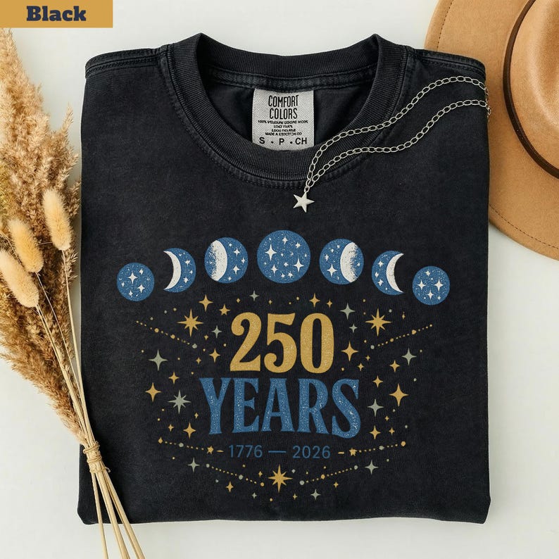 Op de afbeelding: Zwart T-shirt met een hemels ontwerp met maanfasen en sterren. Het shirt toont de tekst "250 YEARS" met de jaren 1776-2026. Een zilveren ketting met een sterhanger ligt op het shirt.
