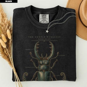 Könnte beinhalten: Ein schwarzes T-Shirt mit einer wissenschaftlichen Illustration eines Hirschkäfers mit dem Text "THE GENTLE DUELLIST" und "Lucanus placidus". Die Grafik enthält Beschriftungen für Mandibel, Antenne, Pronotum und Elytren sowie Farnakzente.